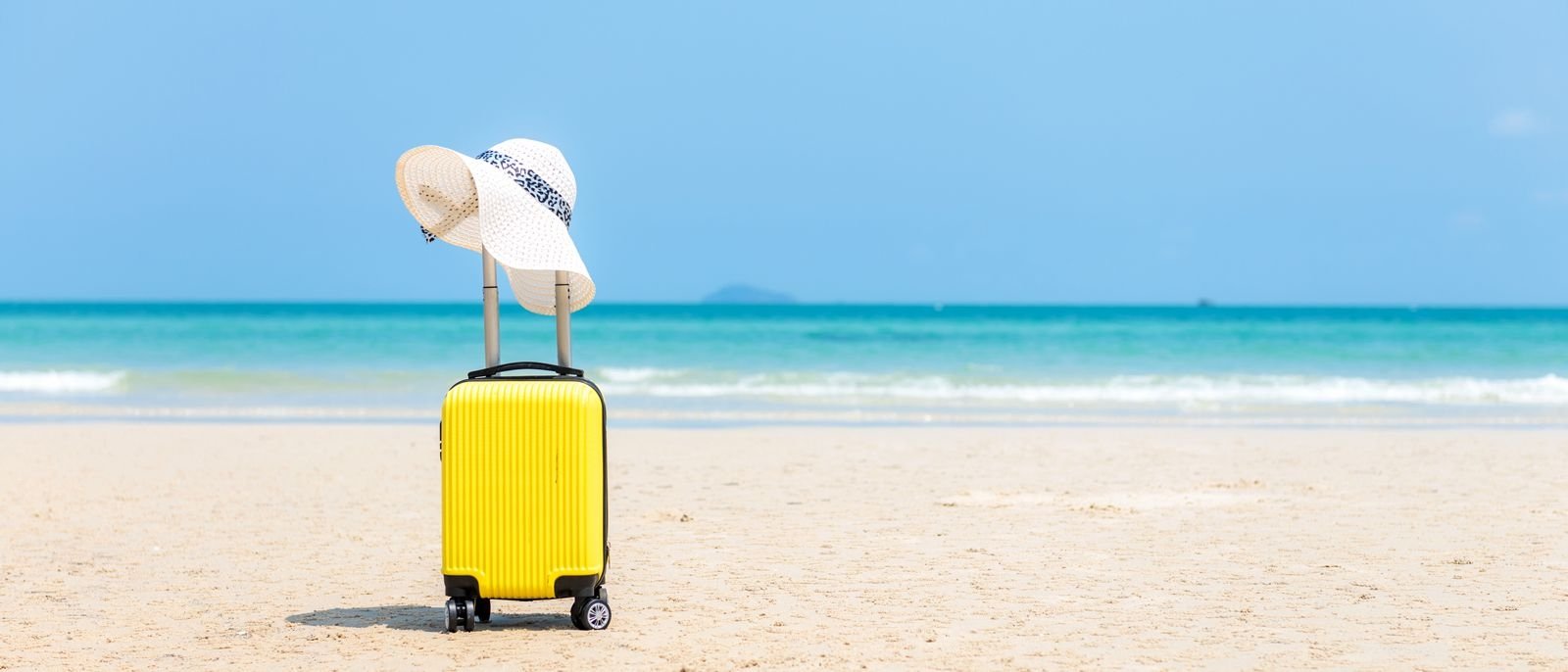 A yellow suitcase on a white sand beach with a floppy sunhat on the handle