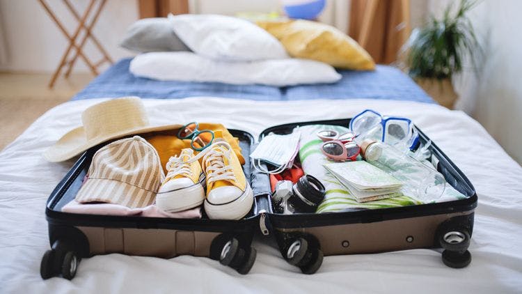 An open suitcase on a bed with neatly packed vacation items including shoes, hats, a camera, and clothing