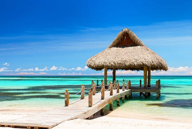 A grass-roofed gazebo at the end of a wooden boardwalk on a white sand beach in Cap Cana