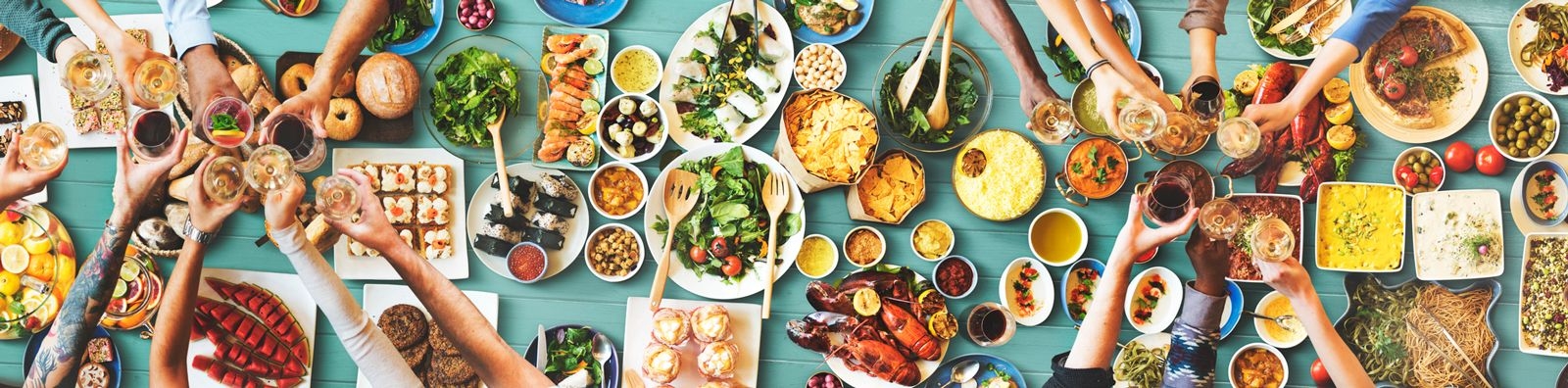 Overhead shot of a light blue table laden with plates of food and people clinking glasses over the top