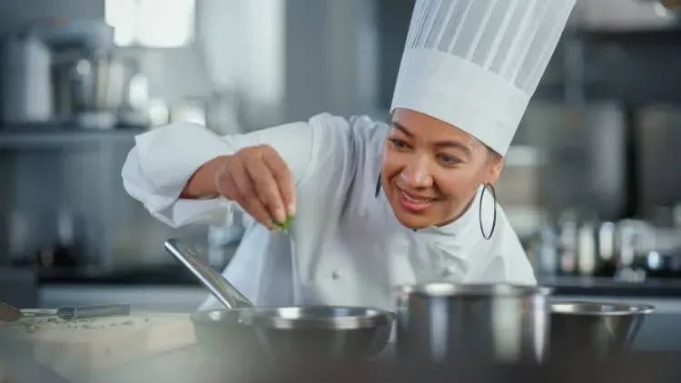 A chef in a whote jacket and tall chef's hat sprinkles salt over a dish that she is preparing