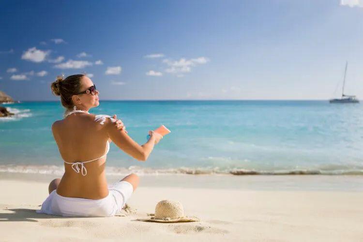 A woman rubs sun cream on her shoulders while sitting on a beach