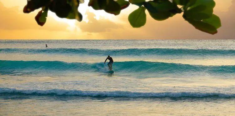 A person stands on a surfboard at sunset, riding a gently rolling wave