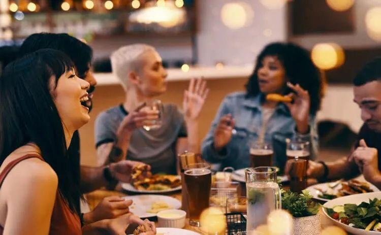 A group of people laughing and talking around a restaurant table with drinks and food
