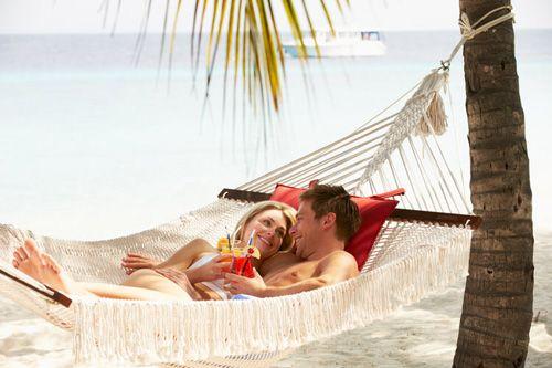 A couple lying in a hammock by the sea drinking cocktails