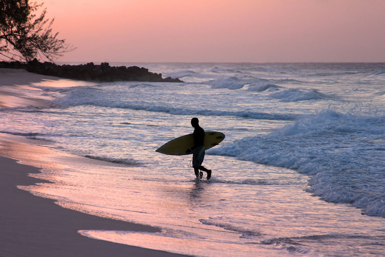 A person carrying a surfboard along the sand at sunrise in Barbados - The best places to visit in June
